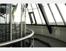 Rundes Innenraum einer modernen Aussichtsplattform mit gläsernen Wänden und Metallgeländern. Große, geneigte Fensterscheiben bieten Ausblick auf eine städtische Umgebung im Sonnenlicht.