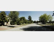 Straße mit Kopfsteinpflaster, flankiert von Bäumen und Gehwegen, rechts ein kleines Gebäude mit angrenzendem Zaun, links ein größeres Gebäude mit Treppenaufgang. Der Himmel ist klar und blau.