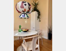 Essbereich mit einem runden weißen Tisch und Holzstühlen auf einem Holzfußboden. Darüber hängt eine große, bunt gemusterte Papierlampe; im Hintergrund stehen eine Zimmerpflanze auf einem weißen Schrank und eine Vase mit Zweigen.