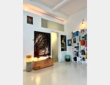 Bright, modern room with white walls and polished light floor, featuring exposed ceiling beams and warm accent lighting. Large abstract paintings hang beside a doorway, and a tall white shelving unit filled with books and objects stands along the right wall.