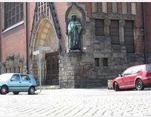 Backsteinfassade eines historischen Gebäudes mit einem gotischen Torbogen und einer Statue in einer Nische. Im Vordergrund eine Pflasterstraße mit geparkten Autos und einem Straßenschild.