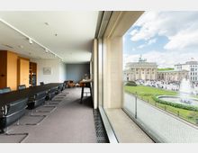 Besprechungsraum mit moderner Einrichtung, darunter ein langer Konferenztisch mit schwarzen Stühlen, daneben eine große Fensterfront mit Blick auf das Brandenburger Tor, einen Springbrunnen und eine belebte Platzanlage.