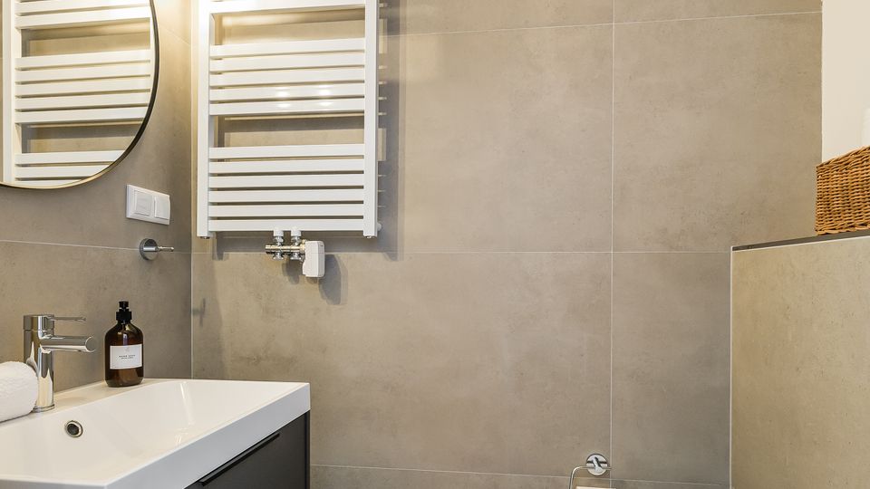Modernes Badezimmer mit großformatigen grauen Fliesen, Waschtisch mit schwarzem Unterschrank und rundem Wandspiegel mit Leuchten. An der Wand hängen ein weißer Handtuchheizkörper und eine Toilette; Einbaustrahler befinden sich in der Decke.