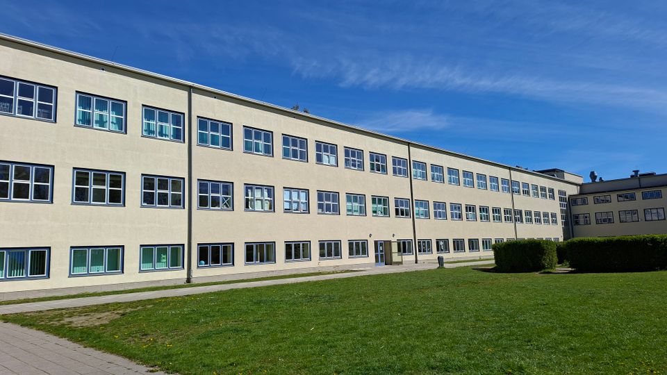 Langer, dreigeschossiger Gebäudetrakt mit heller Fassade und vielen rechteckigen Fenstern in Reihen, davor eine große Rasenfläche und ein Gehweg. Rechts schließen weitere Gebäudeteile sowie geschnittene Hecken an, darüber ein klarer blauer Himmel.