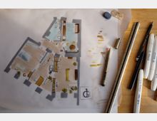 Handgezeichneter Grundriss einer Wohnung auf Transparentpapier mit farbigen Markierungen für Räume und Möbel. Daneben liegen Zeichenwerkzeuge wie Lineal, Stifte und Marker auf einer Holzarbeitsfläche.