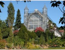 Historisches Gewächshaus aus Glas und Stahl mit spitz zulaufenden Türmen, umgeben von einer vielfältigen Gartenanlage mit Bäumen, Sträuchern und bunten Blumen vor blauem Himmel.