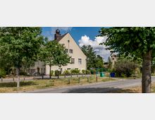 Ein beigefarbenes Gebäude mit Satteldach und einem kleinen Glockenturm im Dachfirst, umgeben von Bäumen und Sträuchern. Im Vordergrund verläuft eine Straße mit Gehweg, daneben stehen mehrere Poller, Fahrräder und eine Mülltonne.