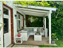 Überdachte Terrasse mit heller Holzstruktur, ausgestattet mit einer weißen Sitzgarnitur und bunten Dekokissen. Die Umgebung wird von dichten grünen Pflanzen eingerahmt, und die Terrasse ist mit Lampen und Blumendekorationen geschmückt.