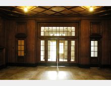 Ein Innenraum mit dunkler Holzvertäfelung, einer Holztür mit Glaseinsätzen in der Mitte und jeweils einer weiteren schmaleren Tür auf beiden Seiten. Die Decke ist mit einem geometrischen Muster gestaltet, und Tageslicht fällt durch die Glastüren herein. Boden und Wände sind mit Stein und Holz gestaltet.