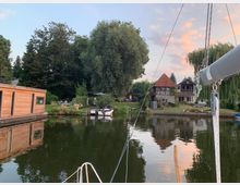 Häuser am Ufer eines kleinen Sees mit Bootsstegen, einem modernen Holzgebäude links, und weiteren Wohngebäuden mit Gärten im Hintergrund. Davor spiegeln sich Bäume und Gebäude im ruhigen Wasser, während im Vordergrund ein Segelboot angeschnitten sichtbar ist.