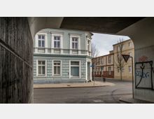 Straßenszene mit einer blauen, historischen Fassade eines zweistöckigen Wohngebäudes im Vordergrund, eingerahmt von einem Betonbogen. Im Hintergrund sind weitere Gebäude mit gelber und roter Klinkerfassade sowie ein Gehweg mit Bäumen zu sehen.