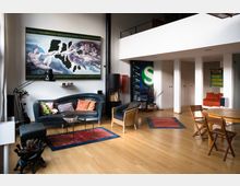 Modernes Wohnzimmer mit einem hellen Holzboden, einer dunkelgrünen Sofagarnitur, einem Glastisch und dekorativen Kissen. An der Wand hängt ein großflächiges Gemälde, daneben ein freistehender Kamin; der Raum wirkt offen mit hohen Decken und einem angrenzenden Essbereich.