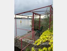 Überwucherter Uferplatz an einem See mit einem roten Metallgerüst über einem kleinen Holzdeck und Geländer. Im Hintergrund Wasserfläche mit Boje und gegenüberliegender Uferbebauung unter bewölktem Himmel.
