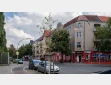 Eine städtische Straßenansicht mit mehrstöckigen Wohngebäuden im Altbaustil, teils mit Balkonen und roten Dächern. Im Erdgeschoss befindet sich ein Restaurant mit dem Namen „Pizzeria“, am Straßenrand stehen parkende Autos, und junge Bäume säumen den Gehweg.