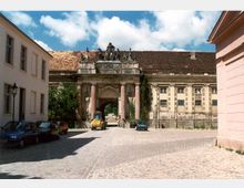 Historisches Tor mit Säulen und verzierter Skulpturengruppe auf einem gepflasterten Platz, umgeben von alten Gebäuden mit beschädigten Fassaden. Im Hintergrund ist ein Durchgang zu sehen, vorne parken mehrere Autos.