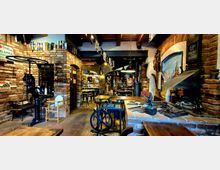 Rustikaler Gastraum mit Backsteinwänden und Holzbalkendecke, eingerichtet mit Holz- und Metalltischen sowie Dekorationen wie einer alten Handpresse und schmiedeeisernen Geräten. Im Hintergrund führt ein gemauerter Rundbogen zu einer Bar mit Regalen und hängenden Lampen.