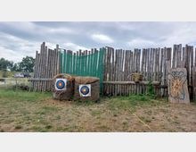 Ein Schießstand im Freien mit zwei Strohbündeln, die Zielscheiben mit farbigen Ringen tragen, vor einer Holzlattenwand und einer grünen Stoffplane. Rechts befindet sich ein Holzbogen mit einem Tierkopf sowie eine Holztafel mit einer bemalten Darstellung.