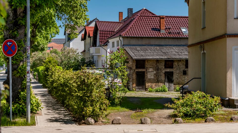 Wohnstraße mit schmalem Gehweg entlang großer Straßenbäume und Hecken, daneben ein kleines Grundstück mit altem Backstein-Nebengebäude. Im Hintergrund stehen Wohnhäuser mit roten Ziegeldächern; vorne rechts ist der Eingang eines beigefarbenen Hauses zu sehen.