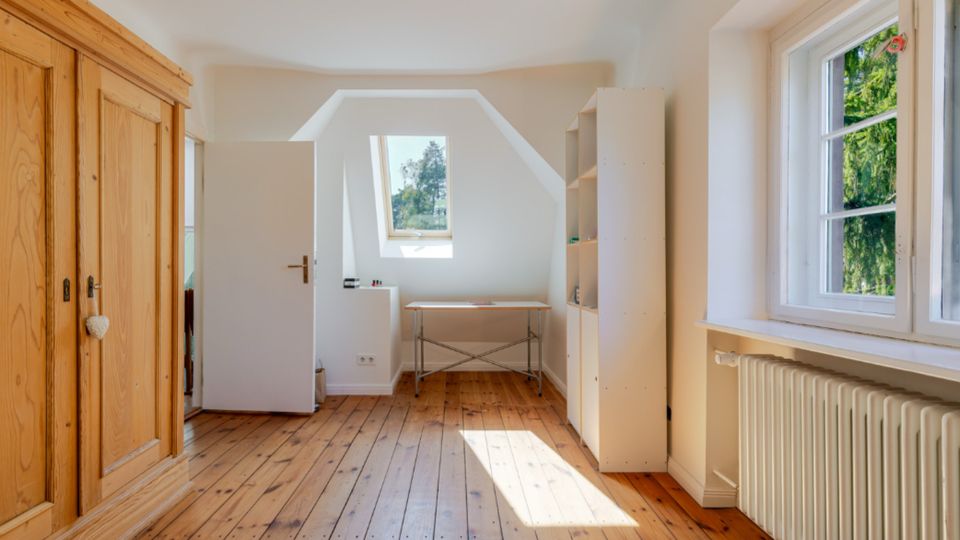 Heller Dachgeschossraum mit Holz-Dielenboden, großem Holzkleiderschrank und einem Schreibtisch unter einem Dachfenster. Rechts sorgen ein breites Seitenfenster mit Blick ins Grüne, ein Heizkörper und ein weißes Regal für eine schlichte Einrichtung.