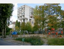 Hochhaus auf Stelzen mit Balkonen im Hintergrund, davor ein öffentlicher Spielplatz mit Klettergerüst, Schaukeln und Bäumen in herbstlicher Umgebung. Im Vordergrund sind Sandflächen und ein Grünstreifen erkennbar.