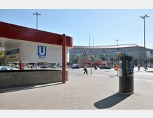 U-Bahn-Eingang mit dem Symbol „U“ und den Linien U3 und U7 am Fehrbelliner Platz in Berlin, umgeben von einem öffentlichen Platz mit Fußgängern, Autos und einem großen Verwaltungsgebäude im Hintergrund.