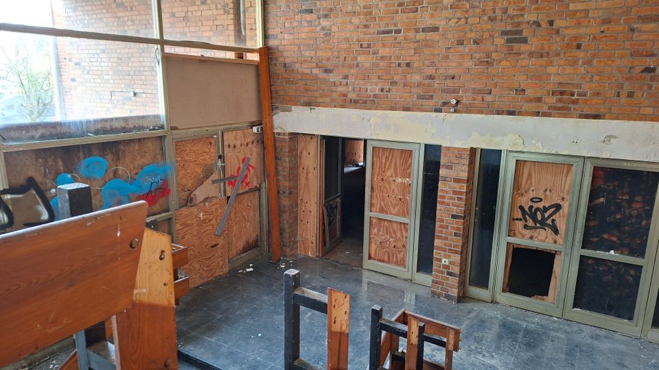 Innenraum eines verlassenen Gebäudes mit hohen Ziegelwänden und großen Fensterflächen; mehrere Türen und Fenster sind mit Sperrholzplatten vernagelt und mit Graffiti beschmiert. Im Vordergrund stehen beschädigte Holzbänke auf einem staubigen Boden.