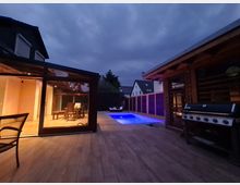 Außenbereich eines modernen Hauses bei Dämmerung, mit einer beleuchteten Veranda, einem Pool mit blauer Beleuchtung, einer überdachten Terrasse und einem Grill. Im Hintergrund sind Nachbarhäuser und hohe Hecken erkennbar.