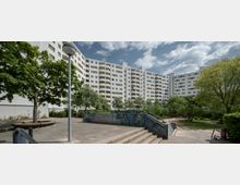 Wohnsiedlung mit weißem, mehrstöckigem Gebäude im Hintergrund, umgeben von Grünflächen, Bäumen und einem gepflasterten Platz mit Betonstufen. Im Vordergrund eine mit Graffiti bemalte Mauer sowie eine Straßenlaterne.