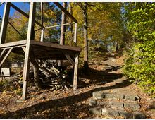 Holzplattform in einem herbstlichen Wald, daneben führt eine steinerne Treppe durch Laub und Bäume hinauf. Die Umgebung ist geprägt von gelb-grünem Laub und vereinzelten Sonnenstrahlen.