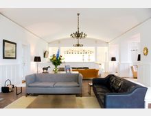 Heller Wohnbereich mit eleganter Einrichtung, einschließlich zweier Sofas, einem Tisch, einem Kronleuchter und weißen Wänden mit Holzvertäfelung. Im Hintergrund stehen Vitrinen mit Pokalen, und der Raum ist mit dekorativen Elementen wie Blumen und Lampen ausgestattet.