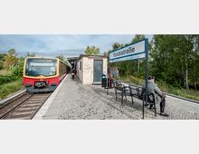 Eine S-Bahn-Station mit dem Namen "Yorckstraße" in Berlin. Links steht ein roter S-Bahn-Zug auf einem Gleis mit der Beschriftung "S25 Teltow Stadt", während rechts Sitzbänke, ein Stationsschild und ein kleines Betriebsgebäude zu sehen sind; im Hintergrund Bäume und Vegetation.