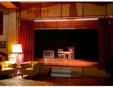 Kleiner Veranstaltungsraum mit einer Bühne, die von roten Vorhängen umrahmt ist. Auf der Bühne stehen ein Holztisch mit zwei Stühlen und ein gepolsterter Stuhl, daneben sind im Vordergrund zwei gelbe Sessel und eine Stehlampe.