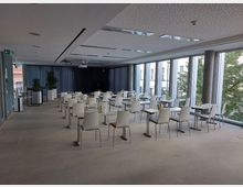 Moderner Konferenzraum mit großen bodentiefen Fenstern, die Blick nach draußen auf Bäume und Stadtgebäude bieten. Der Raum ist mit weißen Tischen und Stühlen eingerichtet, auf den Tischen stehen Getränke und Gläser, dunkle Vorhänge und Pflanzen runden das Bild ab.