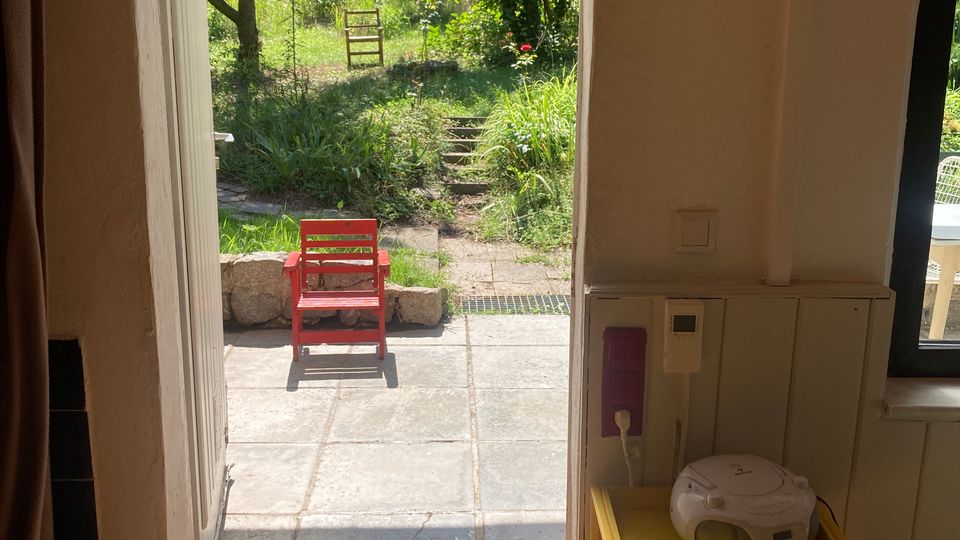 Blick aus einem Innenraum durch eine offene Tür auf eine geflieste Terrasse mit rotem Stuhl; dahinter führt ein schmaler Pfad mit Stufen in einen grünen Garten mit Bäumen und Zaun. Im Vordergrund sind Holzboden und rechts ein kleiner gelber Beistelltisch mit Radio zu sehen.