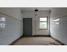 Ein leerer Raum mit weißen Fliesen an den Wänden, einer grauen Tür auf der linken Seite und einem Fenster auf der rechten Seite, das Tageslicht hereinlässt. Der Boden ist unbehandelt und zeigt Spuren von Abnutzung.