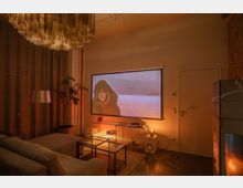 Gemütliches Wohnzimmer mit Heimkino-Setup, bestehend aus einer Leinwand, auf der ein Film projiziert wird. Der Raum hat eine warme Beleuchtung, einen Polstersessel, einen Couchtisch aus Glas und einen Ofen auf der rechten Seite.