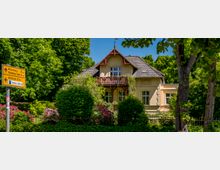 Ein zweigeschossiges, helles Landhaus mit Satteldach und Holzbalkon, umgeben von dichtem Grün, Hecken und blühenden Sträuchern hinter einem niedrigen Zaun. Links im Vordergrund steht ein gelbes Wegweiserschild am Straßenrand.