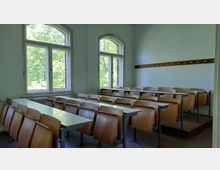 Ein einfacher Klassenraum mit gestuften Sitzplätzen aus Holz und integrierten Tischen, großen Fenstern mit Blick auf grüne Bäume und einer Wand mit einer Garderobenleiste.