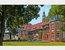 Backsteingebäude im Stil des Historismus mit roten Ziegeldächern, Fachwerkelementen und einem Turm mit Uhr, umgeben von Bäumen und einem gepflegten Rasenbereich, auf dem Parkbänke und Spaziergänger zu sehen sind.