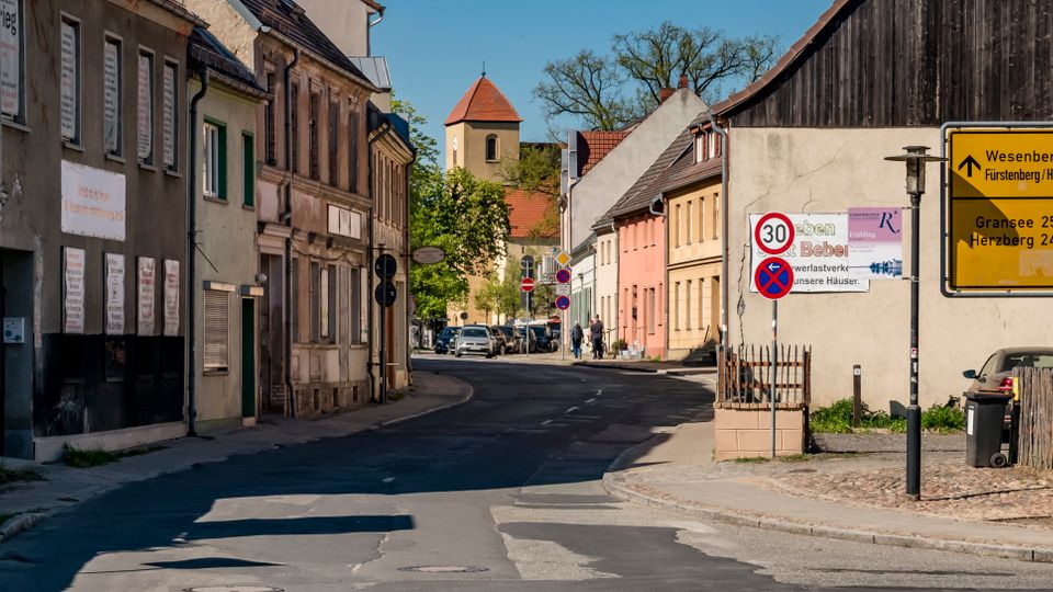 Quiet small-town street lined with older stucco and brick houses, curving toward a church tower with a red roof in the distance. A large yellow German direction sign and speed limit sign stand beside a wooden fence and barn-like building on the right.
