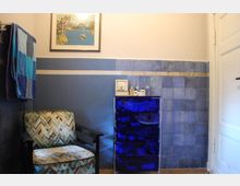 Ein kleiner Raum mit blauen Wandfliesen und einer cremefarbenen oberen Wandfläche. Links steht ein gepolsterter Stuhl mit gemustertem Stoffbezug, daneben ein transparenter blauer Schrank mit mehreren Fächern. Darüber hängen ein gerahmtes Kunstbild und kleinere Pflegeprodukte.