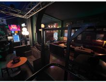 Innenansicht einer dunkel gestalteten Bar mit Holzvertäfelung, Nischen-Sitzbänken und kleinen Tischen entlang der Wände. Links stehen Spielautomaten und eine Lichtanlage mit Metallträgern, während warme Kugelleuchten und gerahmte Wandbilder den Raum beleuchten.