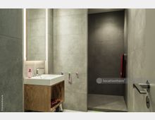 Modernes Badezimmer mit grauen Wandfliesen, einem Waschbecken mit Holzunterschrank und eingebautem beleuchteten Spiegel. Rechts befindet sich ein offener, gefliester Duschbereich mit rotem Handtuch an einem Handtuchhalter.