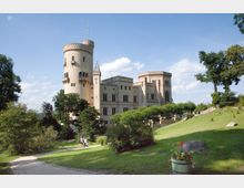 Schloss mit gelblicher Fassade und mehreren Türmen, umgeben von einer gepflegten Parkanlage mit Wiesen, Bäumen und einem Spazierweg; im Vordergrund eine bepflanzte Blumenschale.