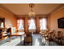 Eleganter Salon- oder Besprechungsraum mit hohen Decken, Stuckrand und zentralem Kronleuchter. Drei große Fenster mit langen Vorhängen lassen viel Tageslicht herein; rechts stehen mehrere gepolsterte Stühle in Reihen, links ein Tisch mit Stuhl sowie ein Bücherregal und ein Schrank.