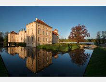 Schlossgebäude im Barockstil mit heller Fassade und roten Dächern, umgeben von einem Wassergraben, der die Spiegelung des Gebäudes zeigt. Im Vordergrund eine kleine Brücke, Wiesenflächen und ein großer Baum mit herbstlich gefärbtem Laub; im Hintergrund ein See und bewaldete Landschaft.