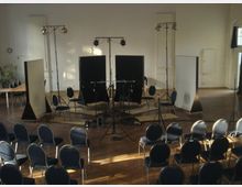 Aufnahme eines Konzertraums oder Studios mit einem runden Stuhlaufbau, Mikrofonstativen und Schallschutzwänden in einem großen, hellen Raum mit Parkettboden. Im Hintergrund befinden sich eine Pflanze, ein Tisch und große Fenster, die Tageslicht hereinlassen.