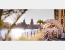 Uferbereich an einem Fluss mit Blick auf die Oberbaumbrücke in Berlin, im Vordergrund eine Außenterrasse mit weißen Möbeln und einem weißen Pavillon, flankiert von einer Palmenpflanze.