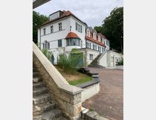 Weiße Villa mit roten Ziegeldächern und grünen Fensterläden, umgeben von Bäumen. Im Vordergrund befinden sich eine Steintreppe und ein gepflasterter Hof mit niedrigen Mauern.