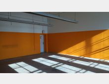 Ein leerer Raum mit einer orangefarbenen Wand und einer weißen Tür, entlang der oberen Wand sind schmale, hängende Leuchten installiert. Durch die große Fensterfront fallen Sonnenstrahlen, die Schattenmuster auf den Boden und die Wand werfen.